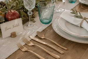 Matte gold flatware beside handmade ceramic dinnerware, linen napkin, and Italian-style place card on a rustic wooden Christmas table.