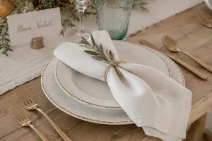 Mediterranean Christmas place setting with handmade ceramic plates, white linen napkin tied with twine and olive branch, and matte gold flatware.